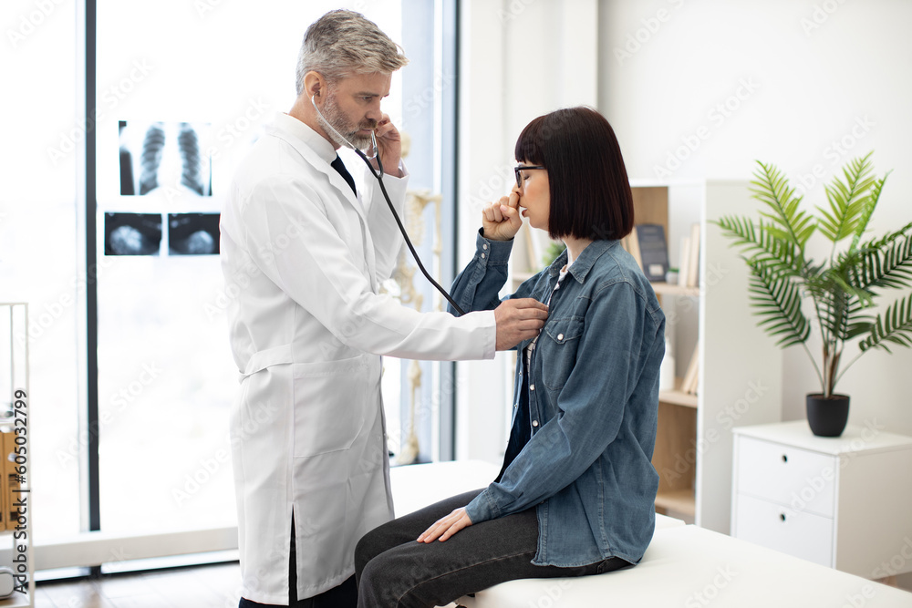 Beautiful woman in jeans outfit coughing while sitting on exam couch during medical checkup in private practice. Focused family doctor listening to lungs of lady with stethoscope in consulting room. - obrazy, fototapety, plakaty 