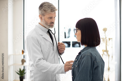 Young caucasian woman in casual wear inhaling when breathing while mature male holding stethoscope on chest. Efficient general practitioner checking lungs of female with doctor's listening device.