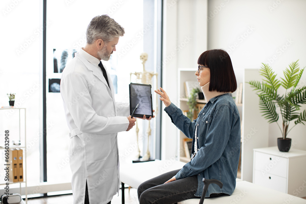 Obraz premium Confident mature man in lab coat holding digital tablet while young lady in cervical collar sitting in exam room. General practitioner showing test results to female with car accident injury.
