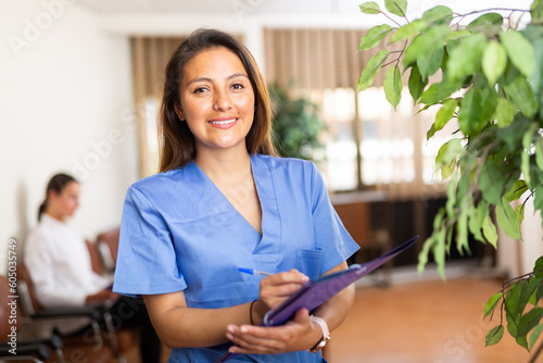 Positive Latino doctor woman standing in office with clipboard, writing medical history sheet