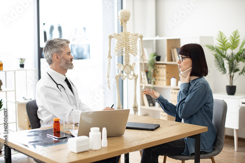 Φωτογραφία Side view of professional man in lab coat and attractive woman in C collar sitting at writing desk in doctor's office