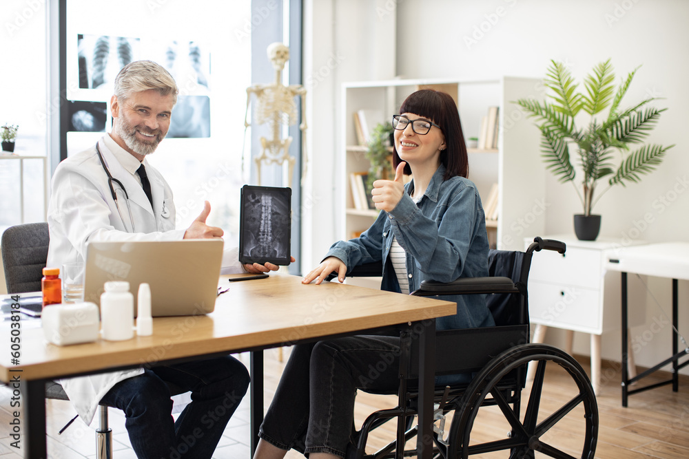 Full length portrait of friendly doctor and happy woman with disability on meeting in consulting room of clinic. Cheerful physician showing x-ray on tablet with positive results to lady in wheelchair. Full length portrait of friendly doctor and happy woman with disability on meeting in consulting room of clinic. Cheerful physician showing x-ray on tablet with positive results to lady in wheelchair.