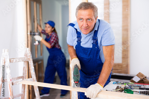 Man builder in blue overall using fretsaw on wooden plank during renovation works in apartment.