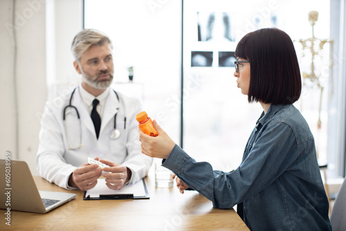 Side view of pretty woman wearing jeans outfit looking at medications in glass container in clnic. Caucasian female in glasses questioning aged male doctor about side effects of pills.
