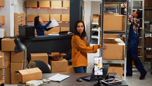 Asian woman doing teleshopping ad for social media promotion, showing merchandise on streaming platform for startup business development. Warehouse owner promoting stock online.