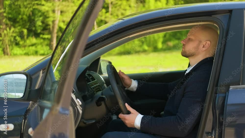 a young adult Caucasian man in a suit is sitting in a car touching the steering wheel with his hands a young adult Caucasian man in a suit is sitting in a car touching the steering wheel with his hands