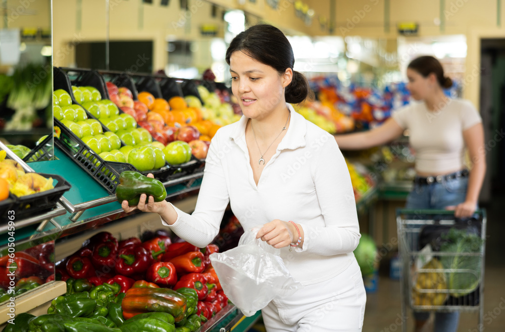 Young woman shopper in casual clothes chooses bell pepper in supermarket