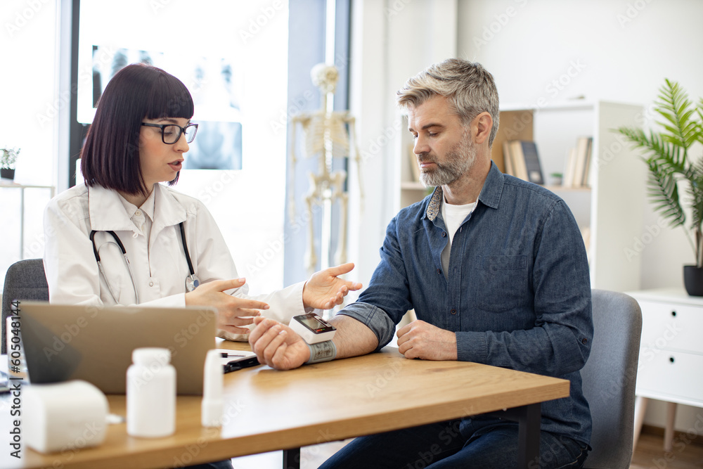 Attractive cardiologist telling patient about all possible reasons of hypertension and keeping medical history on laptop. Adult man listening explanations about necessity of leading healthy lifestyle. Attractive cardiologist telling patient about all possible reasons of hypertension and keeping medical history on laptop. Adult man listening explanations about necessity of leading healthy lifestyle.