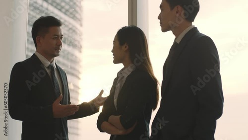 three asian business people colleagues standing and talking by the window in office at dusk