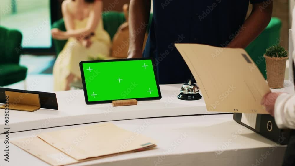 Guest asking about greenscreen info, pointing at tablet on reception counter in hotel lobby. Receptionist talking to traveller about isolated display mockup template, front desk concierge service.