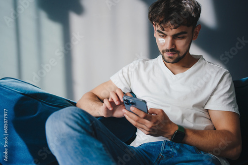 Indoor shot of caucasian young man surfing internet on mobile phone, messaging friends online relaxing on sofa. Guy browsing internet, surfing web