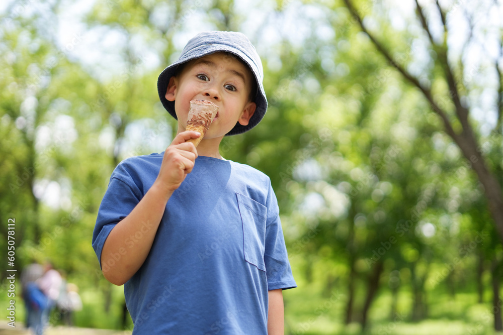 Cute little kid boy in a hat eating ice cream cone standing in a park looking at camera. Summer concept. Eating ice-cream.