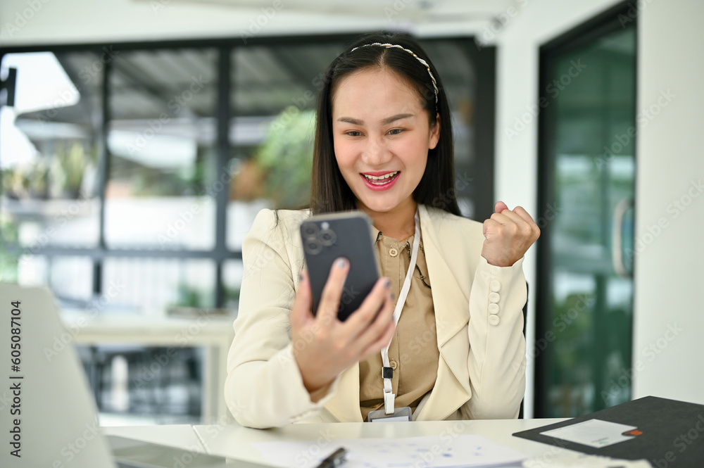 Obraz premium Excited Asian businesswoman showing clenched fist, looking at her smartphone screen