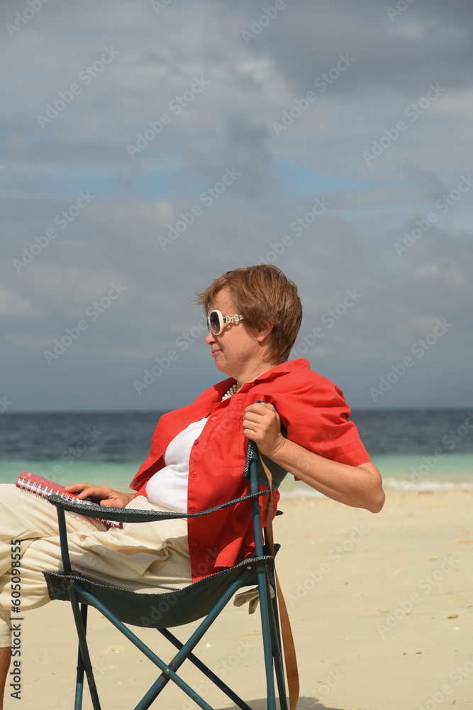 Fototapeta premium Mature woman writer in sunglasses writing in notebook. Senior woman writer is writing details on notebook while working at the beach. Active lifestyle after retirement. Side view