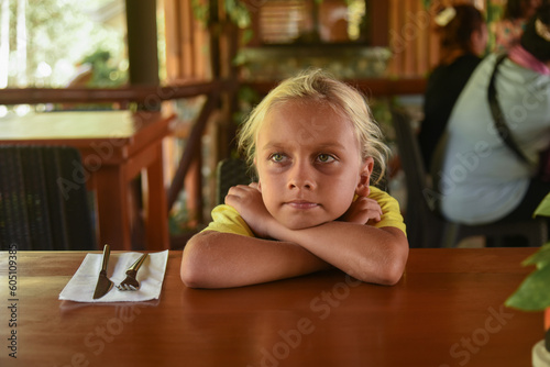 An american blonde boy waiting for a dinner in restaurant. Portrait of child with long blonde hair and a yellow T-shirt sitting in a cafe. Bad appetite and digestive problems on a diet