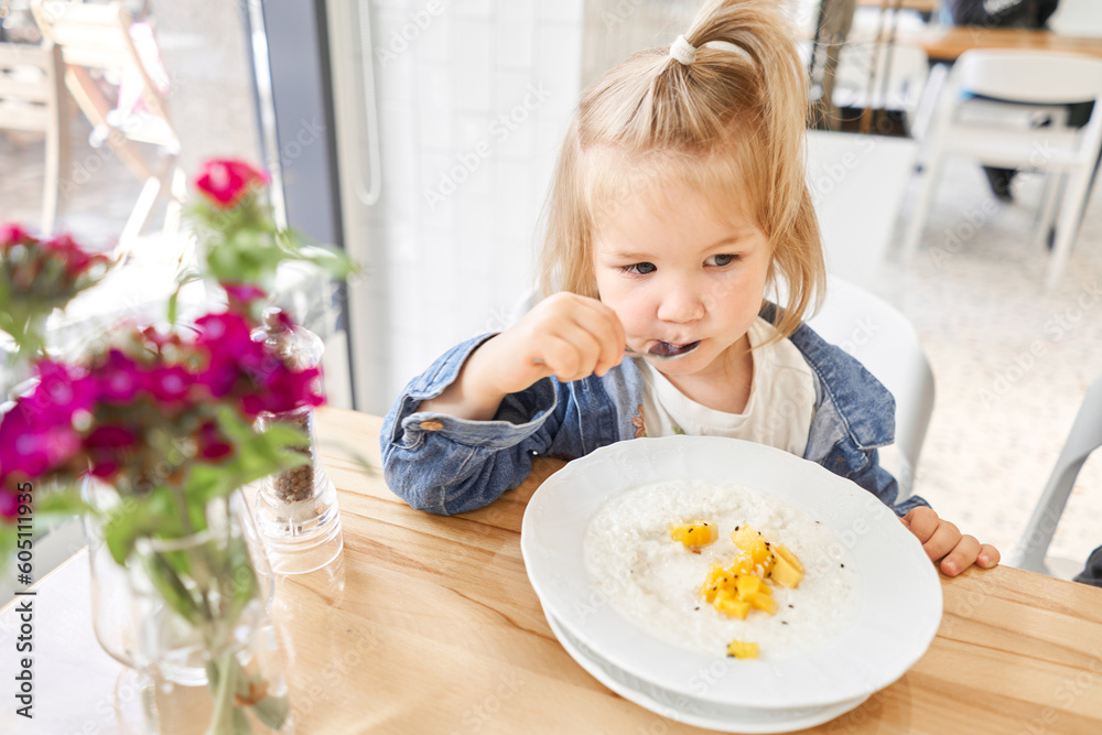 Cute little girl drinking eating rice porridge with mango. enjoying breakfast. Kid having breakfast at cafe. Cute little girl drinking eating rice porridge with mango. enjoying breakfast. Kid having breakfast at cafe.