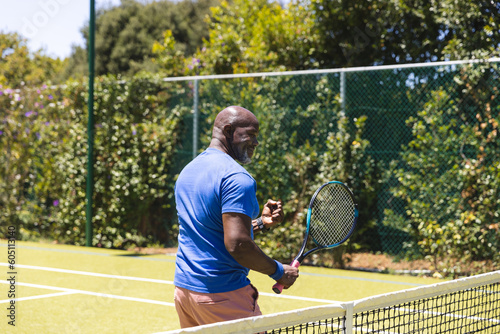 Happy senior african american man playing tennis and celebrating on sunny grass court
