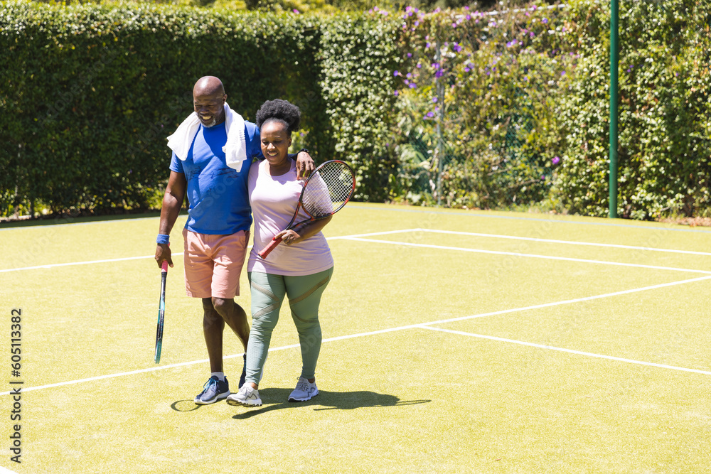 Happy senior african american couple holding tennis rackets walking on sunny grass court, copy space