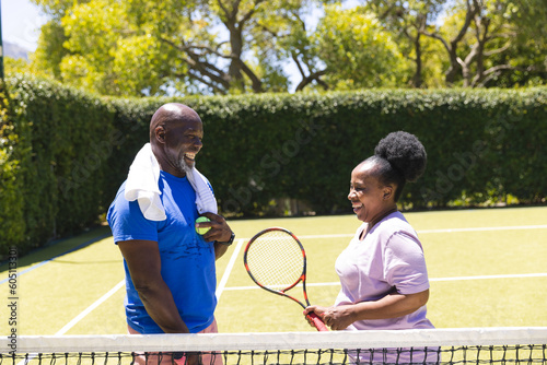 Happy senior african american couple with tennis ball and racket talking on sunny grass court