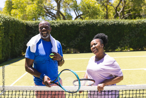 Happy senior african american couple with tennis rackets laughing on sunny grass court