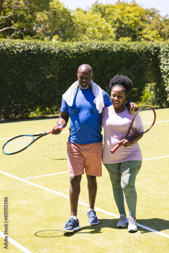 Happy senior african american couple holding tennis rackets and walking on sunny grass court