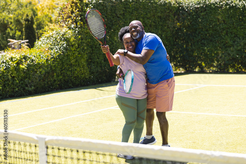 Portrait of happy senior african american couple with rackets embracing on sunny grass tennis court © wavebreak3
