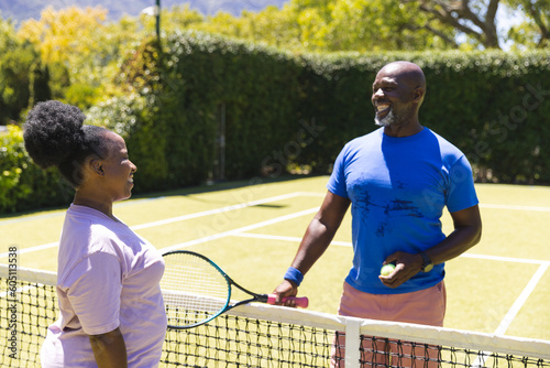Happy senior african american couple with tennis racket talking over net on sunny tennis court © wavebreak3