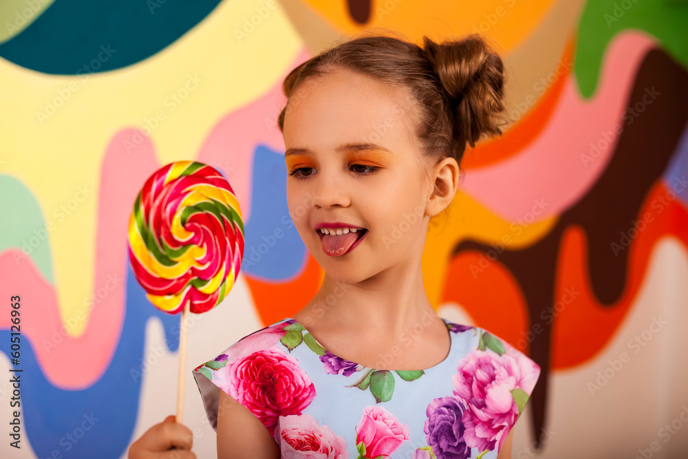 Joyful little girl stuck out her tongue, looking on round lollipop at multi colored wall. Lovely kid 6-7 year old in colorful flowers dress with candy on stick. Summer sweet concept. Copy text space