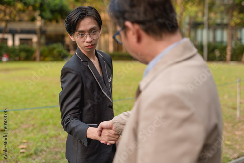 A young asian man is congratulated by his boss for a promotion. Closing a deal with a business partner while walking at the park. A genuine and solid handshake. Outdoor city park scene.