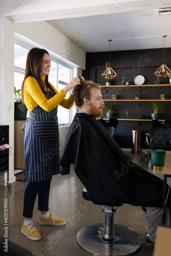 Happy caucasian female hairdresser undoing hair bun of long haired male client at hair salon