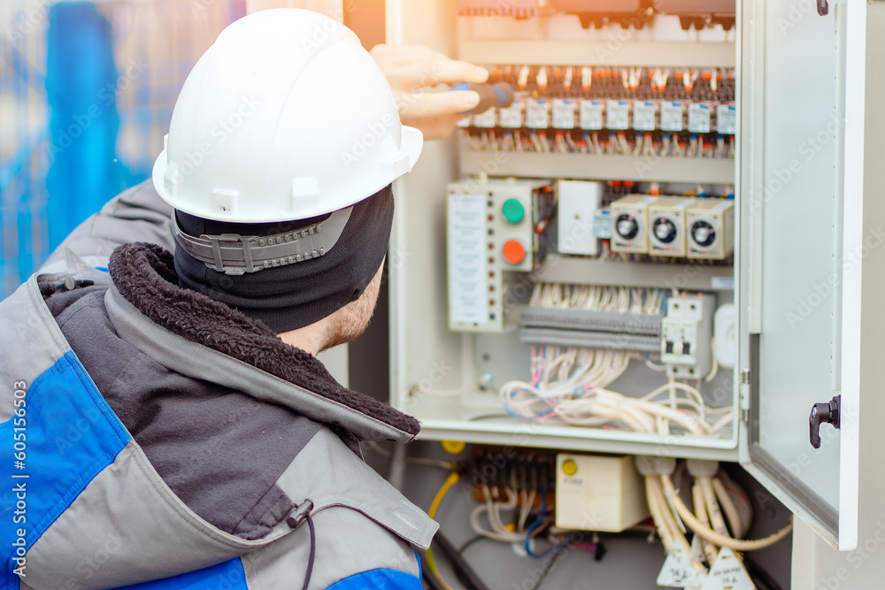 Professional electrician connects wires in electrical panel room outside. Worker in hard hat and winter clothes checks equipment with screwdriver. Back view. - obrazy, fototapety, plakaty 