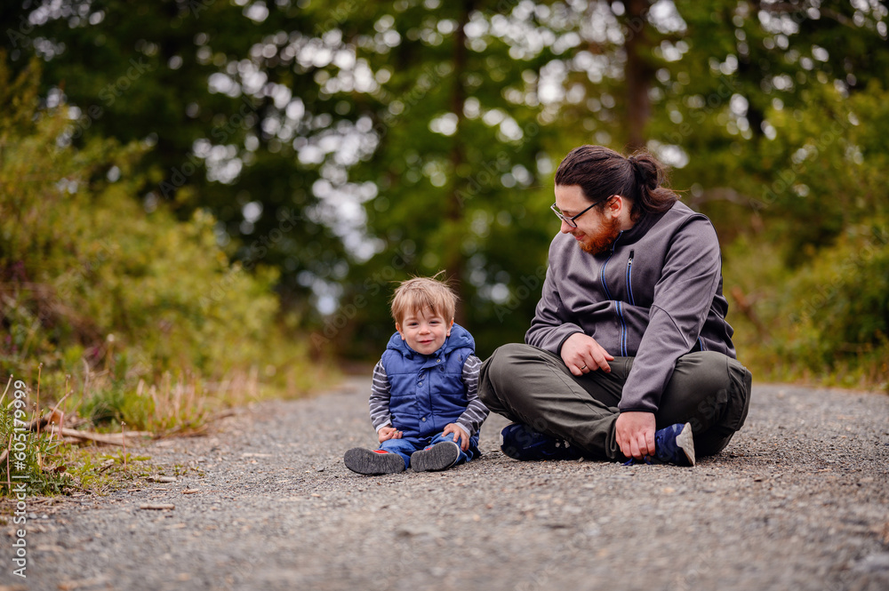 Fototapeta premium Young long hair bearded man in glasses sitting with little toddler boy on road playing with stones