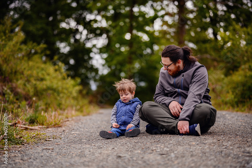 Young long hair bearded man in glasses sitting with little toddler boy on road playing with stones