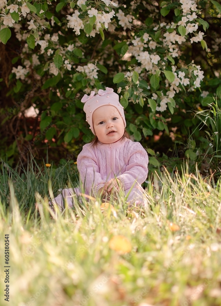 Happy little girl is playing in spring garden. Charming one-year-old girl in pink jumpsuit enjoys walk in fresh air. Child is sitting on green grass. Vertical photography.