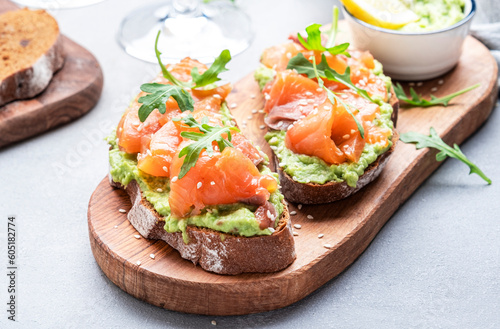 Avocado salmon sandwich on rye bread with guacamole sauce, young arugula and sesame seeds on wooden cutting board, gray table background, top view