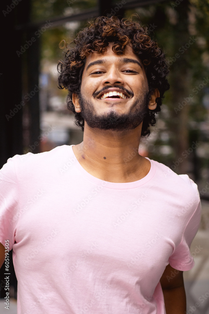 Portrait of young and curly indian man in pink t-shirt looking at camera and smiling while standing on blurred urban street Portrait of young and curly indian man in pink t-shirt looking at camera and smiling while standing on blurred urban street
