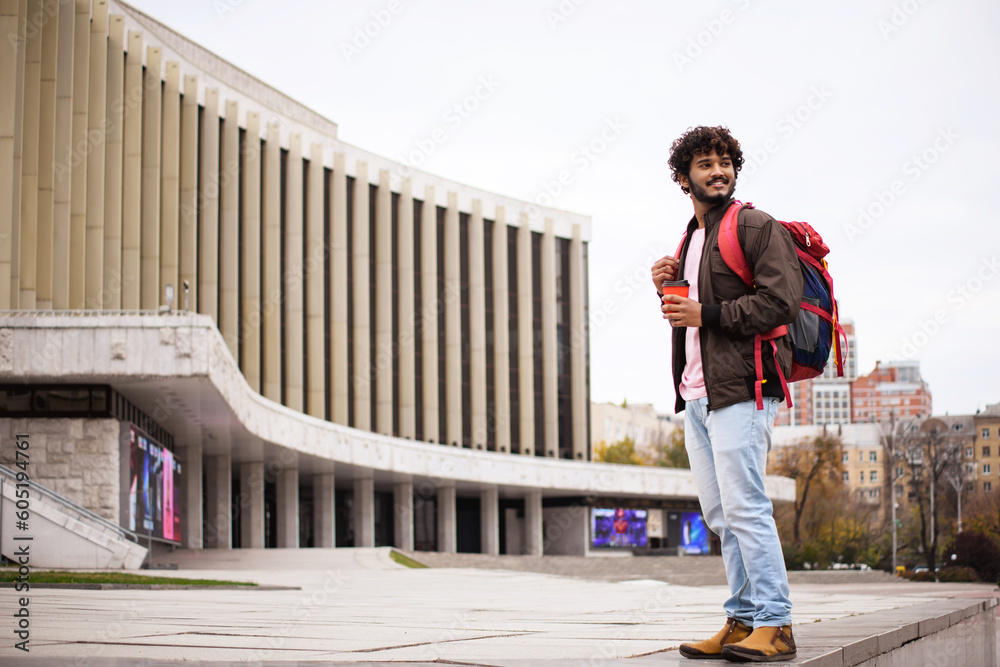 Positive young indian man in casual clothes holding backpack and coffee to go while standing on blurred urban street 