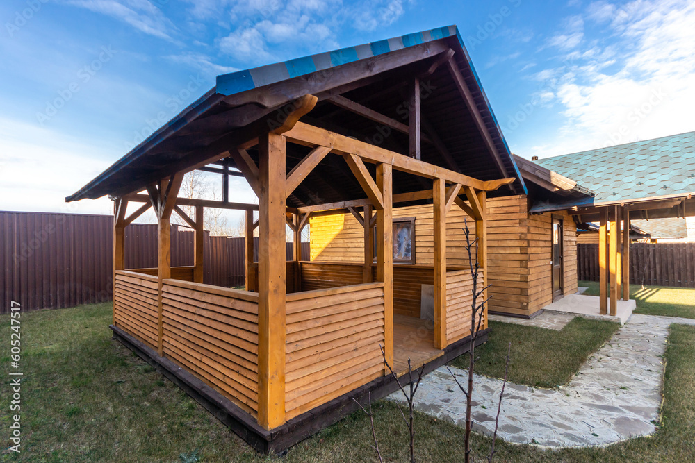 A gazebo made of wood and a bathhouse on a private territory, against the blue sky. A green lawn with stone paths.