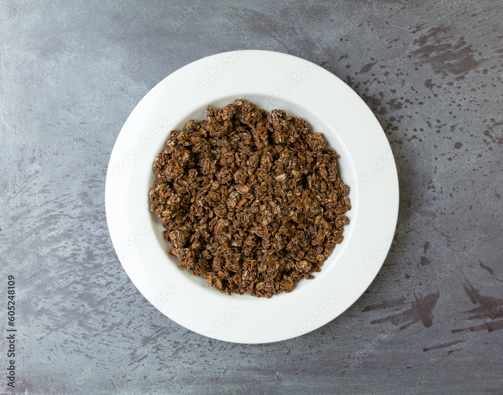 Overhead view of a serving of sweet chocolate granola in a white bowl on a gray tabletop. Overhead view of a serving of sweet chocolate granola in a white bowl on a gray tabletop.
