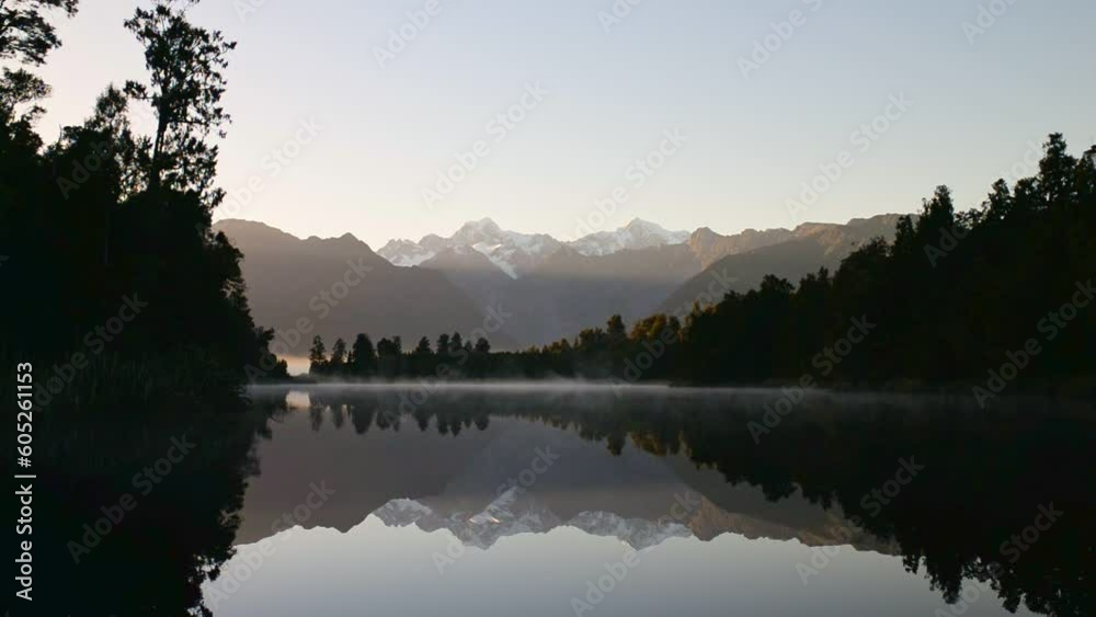 Serene, misty sunrise over Lake Matheson, New Zealand; snowy mountain peaks reflecting in calm water