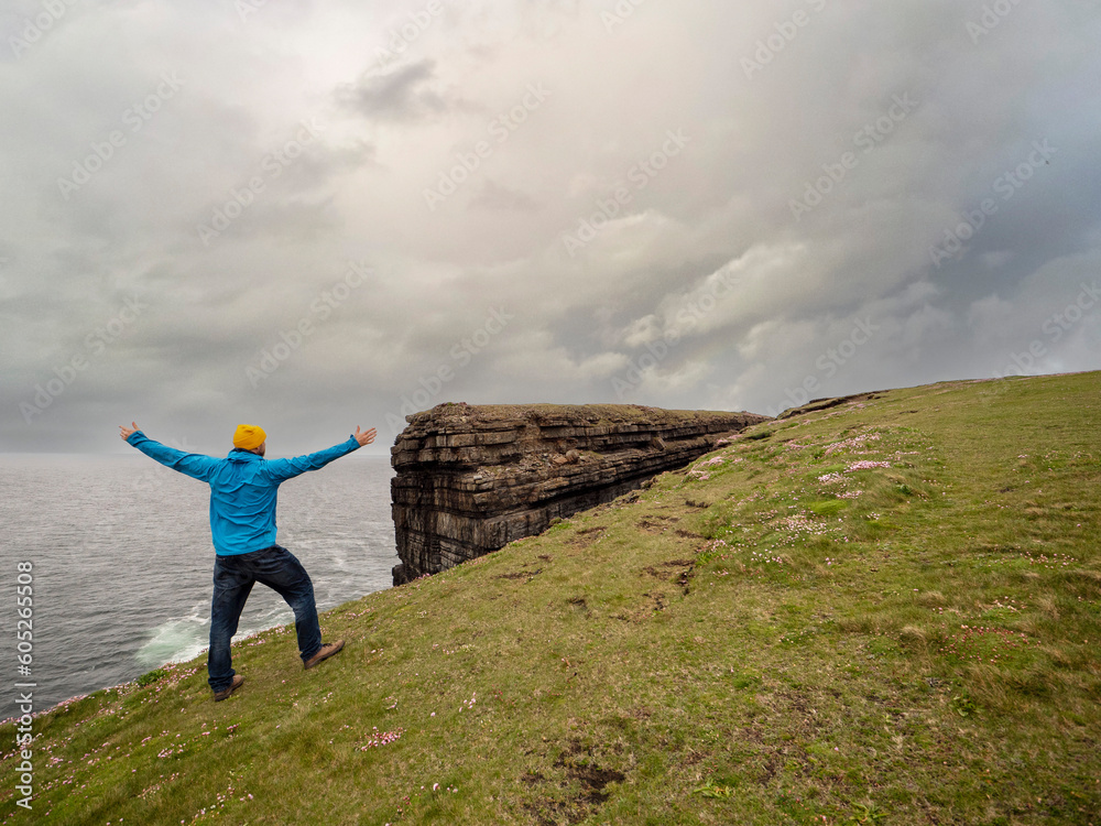 Obraz premium Male tourist in blue jacket and yellow hat standing on edge of a cliff, Loop head area county Clare, Ireland. Dramatic sky. Travel and sightseeing