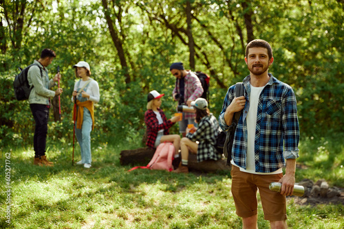 Young happy, smiling man in checkered shirt going hiking with friends. Group of young people walking in forest. Concept of active lifestyle, nature, sport and hobby, friendship, leisure time