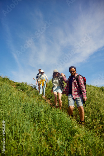 Group of friends, young people walking in meadow, going down the hill on warm, sunny day. Men giving helping hand to women. Concept of active lifestyle, nature, sport and hobby, friendship