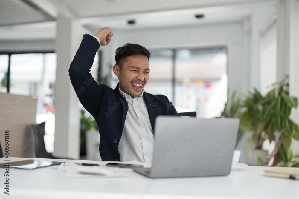 Happy young businessman looking at laptop excited by good news online, lucky successful winner man sitting at office desk raising hand in yes gesture celebrating business success win result Happy young businessman looking at laptop excited by good news online, lucky successful winner man sitting at office desk raising hand in yes gesture celebrating business success win result