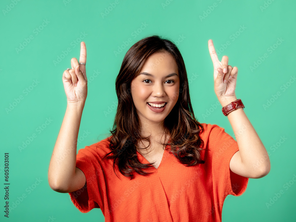 A studio portrait of a beautiful young Asian Indonesian woman wearing an orange long-sleeve shirt pointing up while smiling. Isolated on a green background. A studio portrait of a beautiful young Asian Indonesian woman wearing an orange long-sleeve shirt pointing up while smiling. Isolated on a green background.
