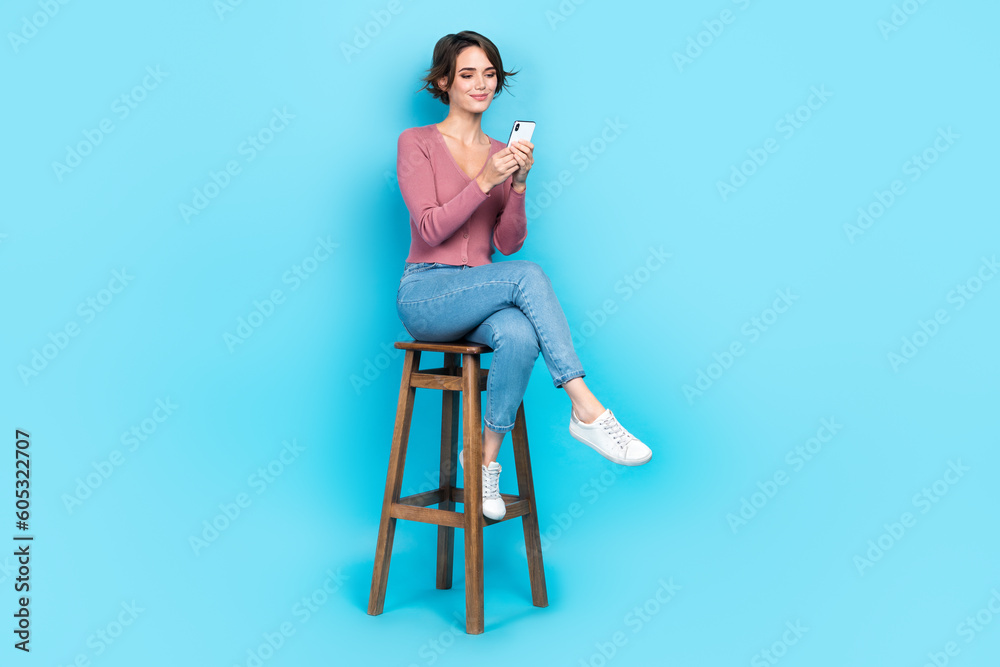 Full length photo of adorable girl wear pink cardigan denim trousers sitting on chair look at smartphone isolated on blue color background Full length photo of adorable girl wear pink cardigan denim trousers sitting on chair look at smartphone isolated on blue color background