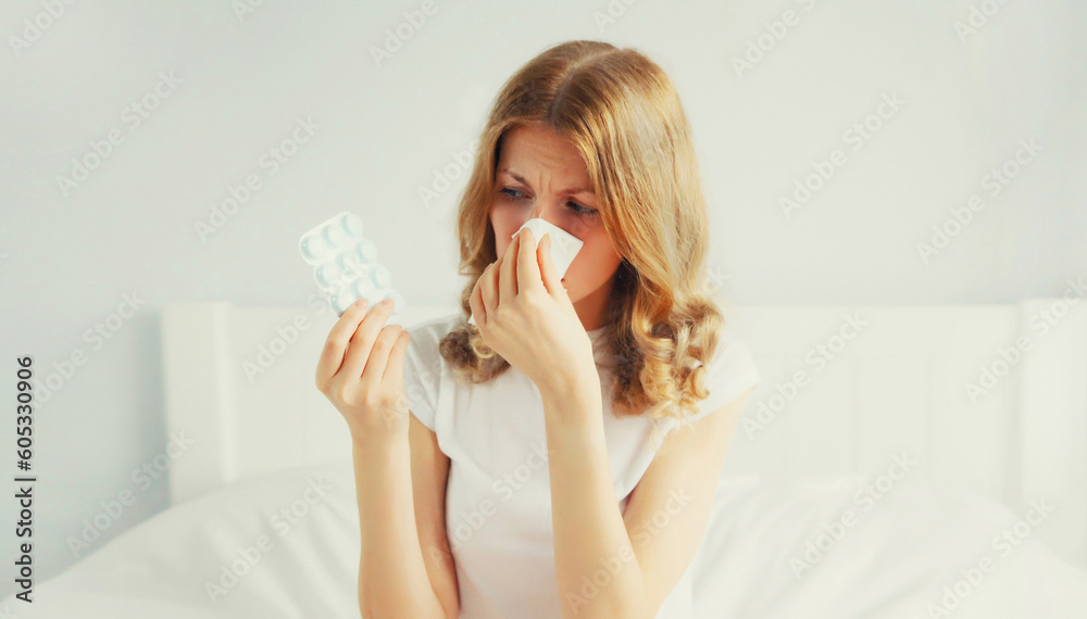 Portrait of sick upset woman sneezing blow nose using tissue and looking at the pills, female patient does not want to take medicine on the bed in bedroom