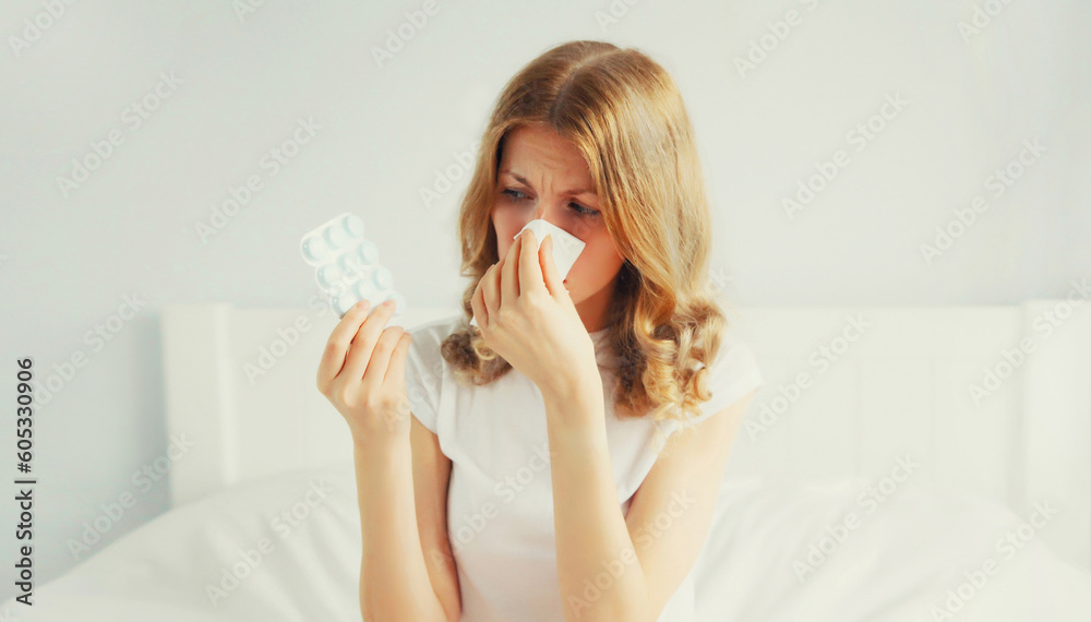 Portrait of sick upset woman sneezing blow nose using tissue and looking at the pills, female patient does not want to take medicine on the bed in bedroom Portrait of sick upset woman sneezing blow nose using tissue and looking at the pills, female patient does not want to take medicine on the bed in bedroom