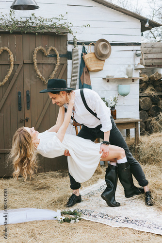 young couple newlyweds bride in short white dress and wreath with veil and groom in hat and jeans with suspenders hugging and having fun at barn of country house
