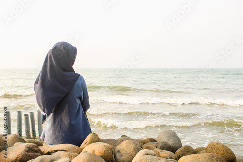 A young woman wearing a hijab sits on a rock overlooking the beach